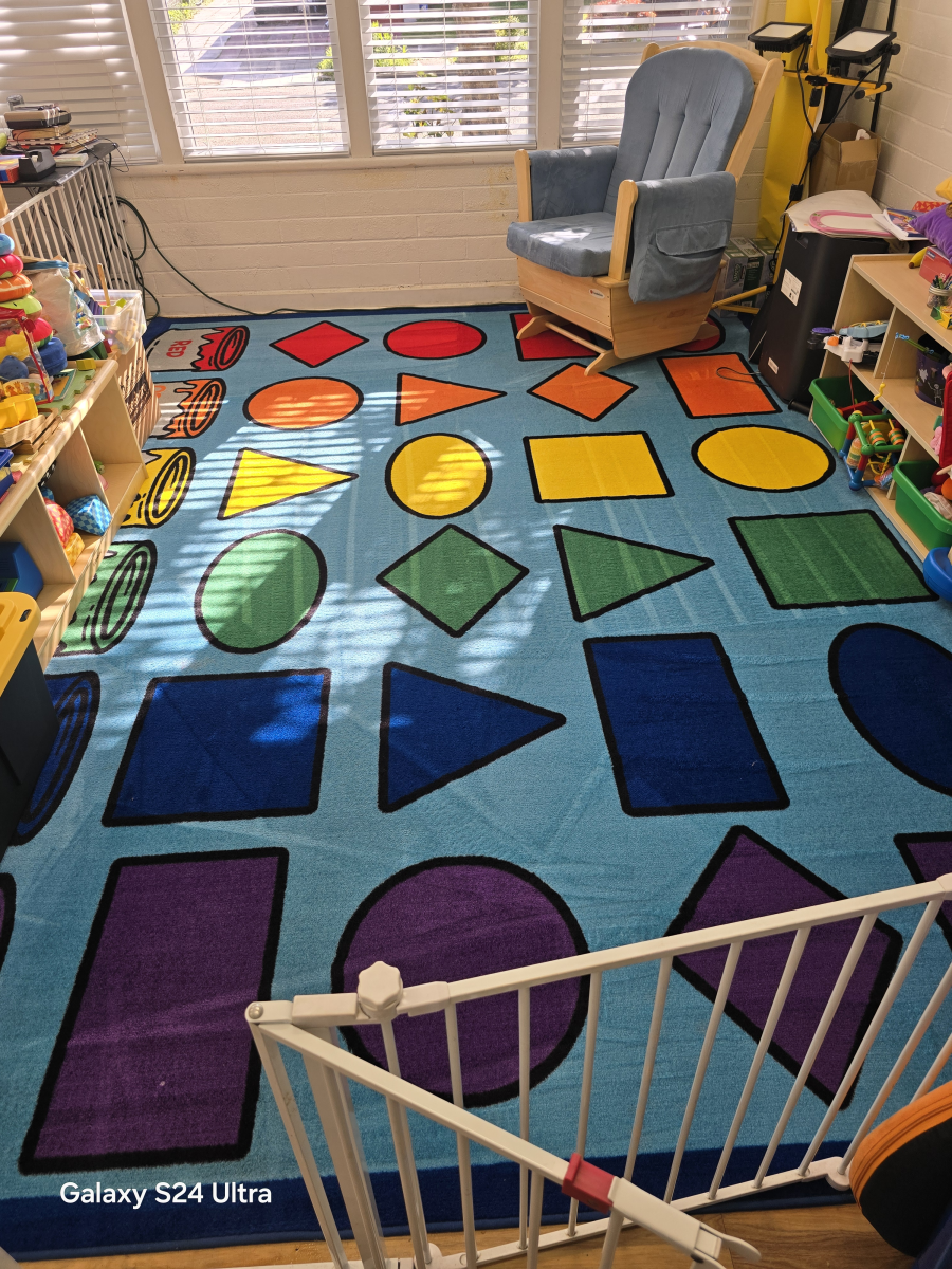 A colorful children's rug featuring various shapes in rows, placed in a room with a light blue rocking chair and toy bins.