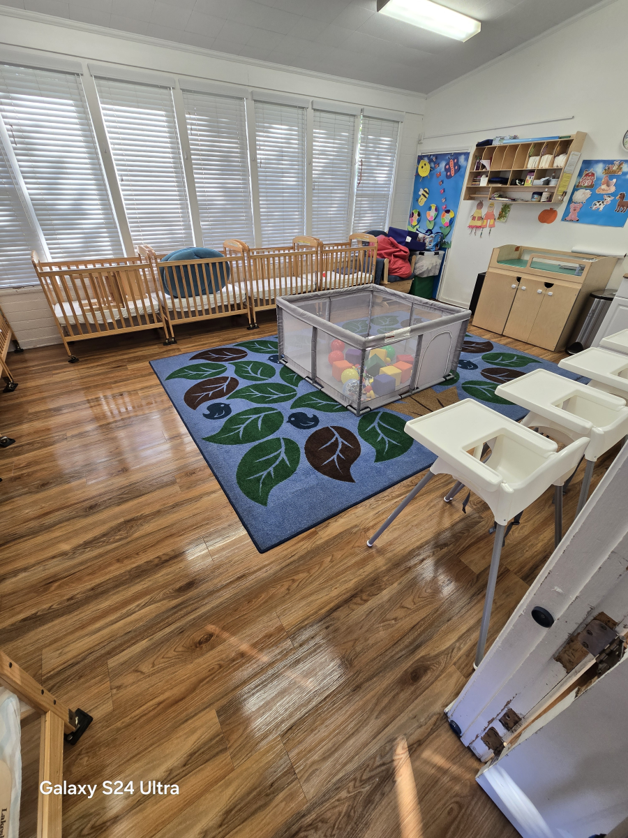 An indoor daycare room with several wooden cribs against a large window, a central play area, and white high chairs.