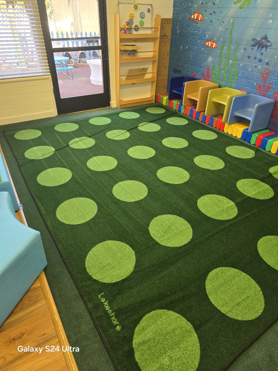 A rectangular green rug with light green circles sits in a playroom with colorful toy chairs and a nautical wall mural.