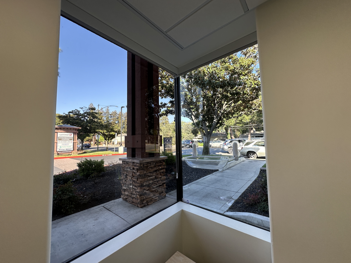 A view through a corner window showing an outdoor sidewalk, landscaping, a stone-based pillar, and a parking lot.