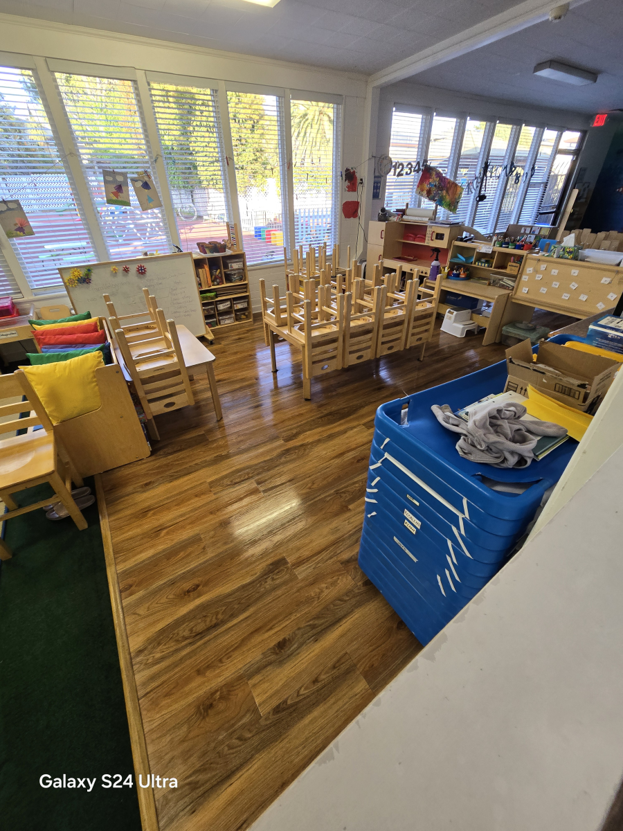 A bright preschool classroom with wooden floors, small chairs, tables, and a large blue storage bin.