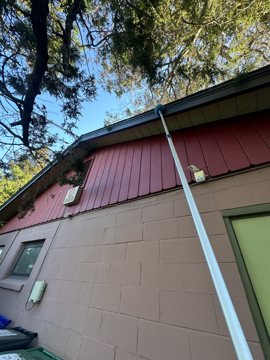 An extendable pole reaches up toward the gutter of a building with a red wood-paneled upper wall and tan block lower wall.