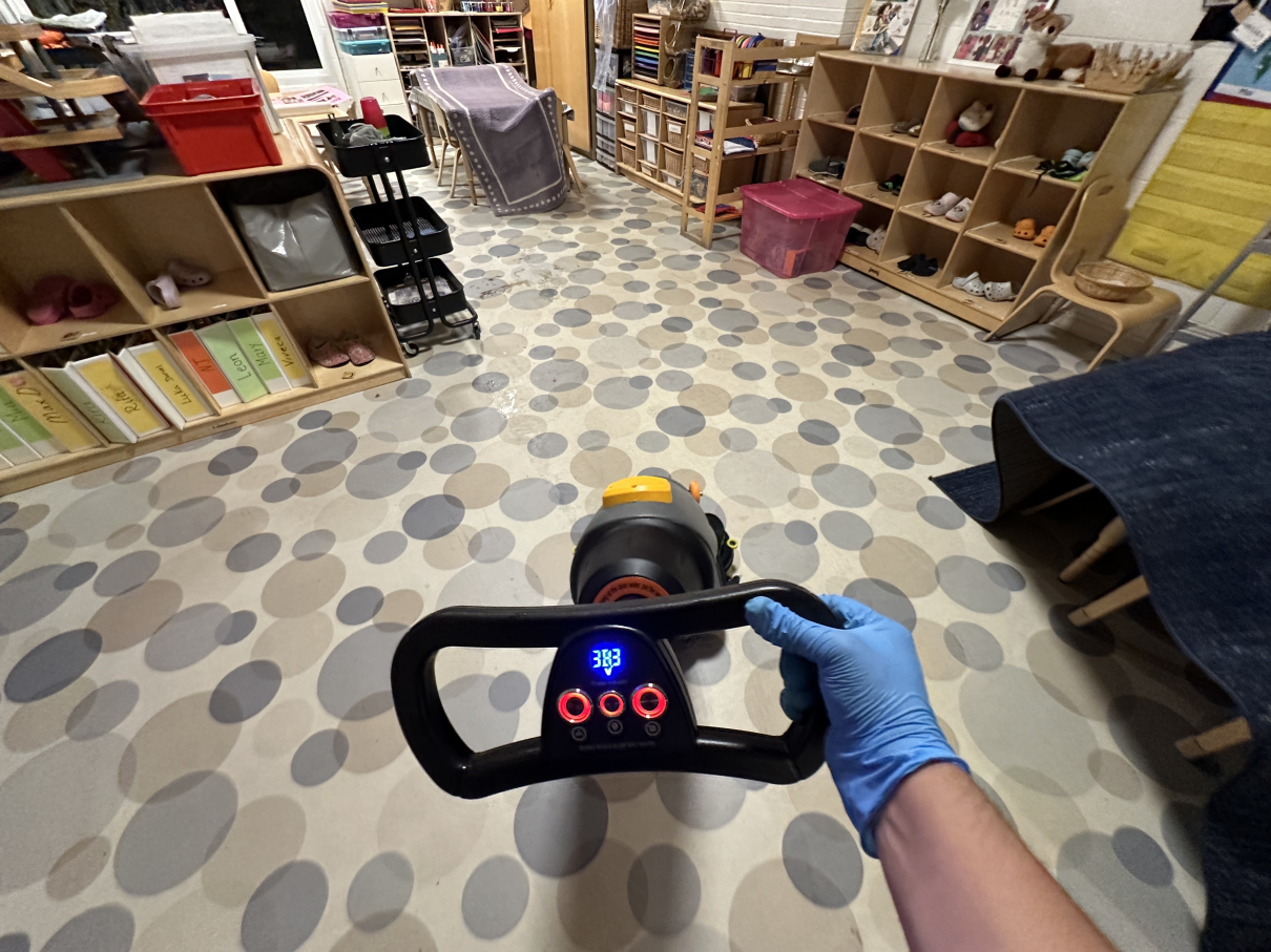 A hand in a blue glove holds the handle of a cleaning machine on a room floor with a polka-dot pattern.