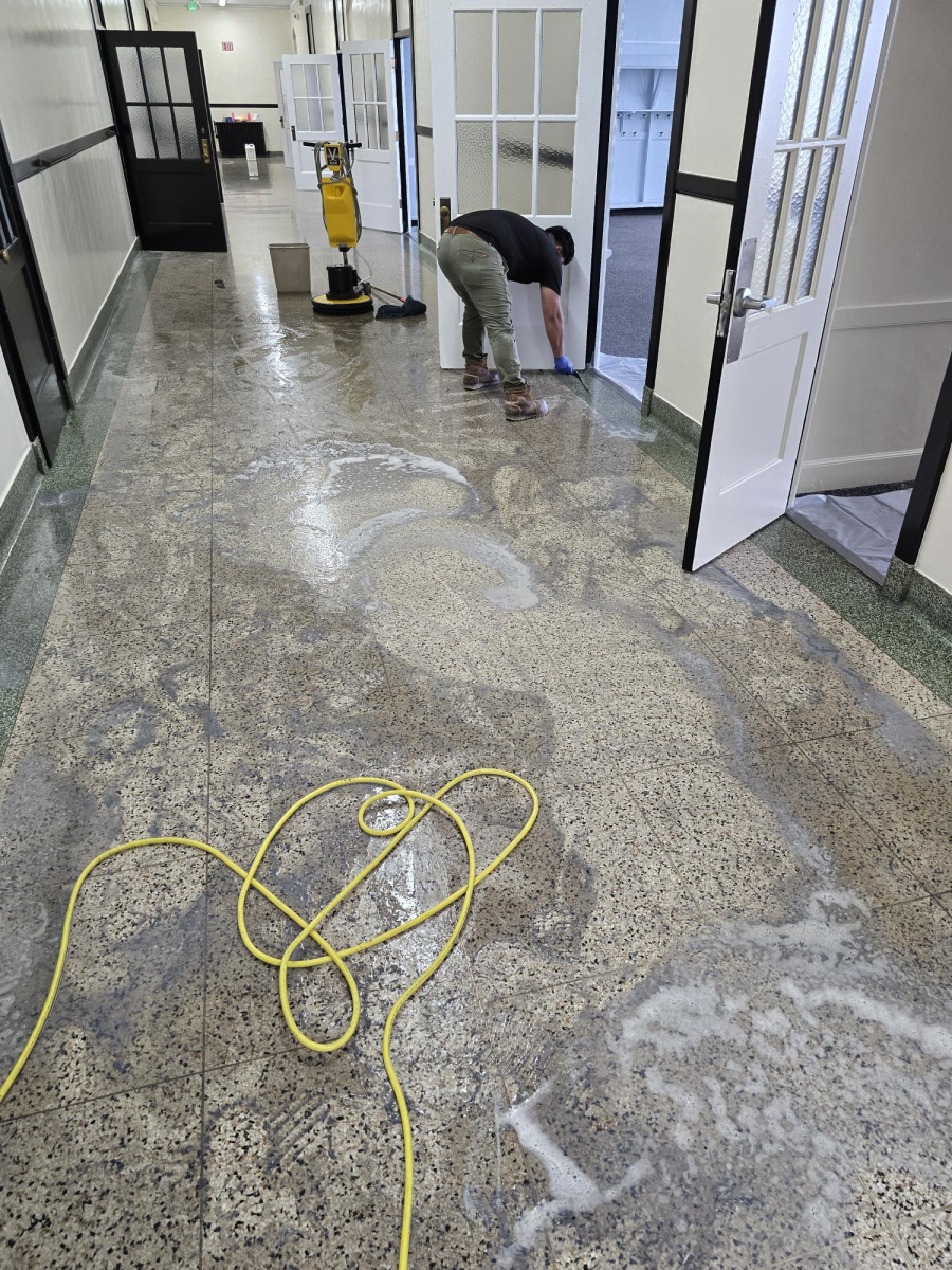A person cleans a speckled floor in a hallway with a yellow hose and a floor buffer nearby.