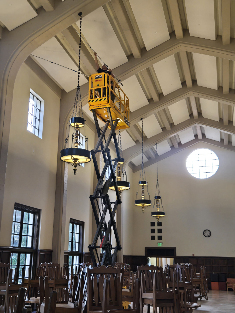 A worker in a yellow scissor lift reaches up to repair a large hanging chandelier in a high-ceilinged room.