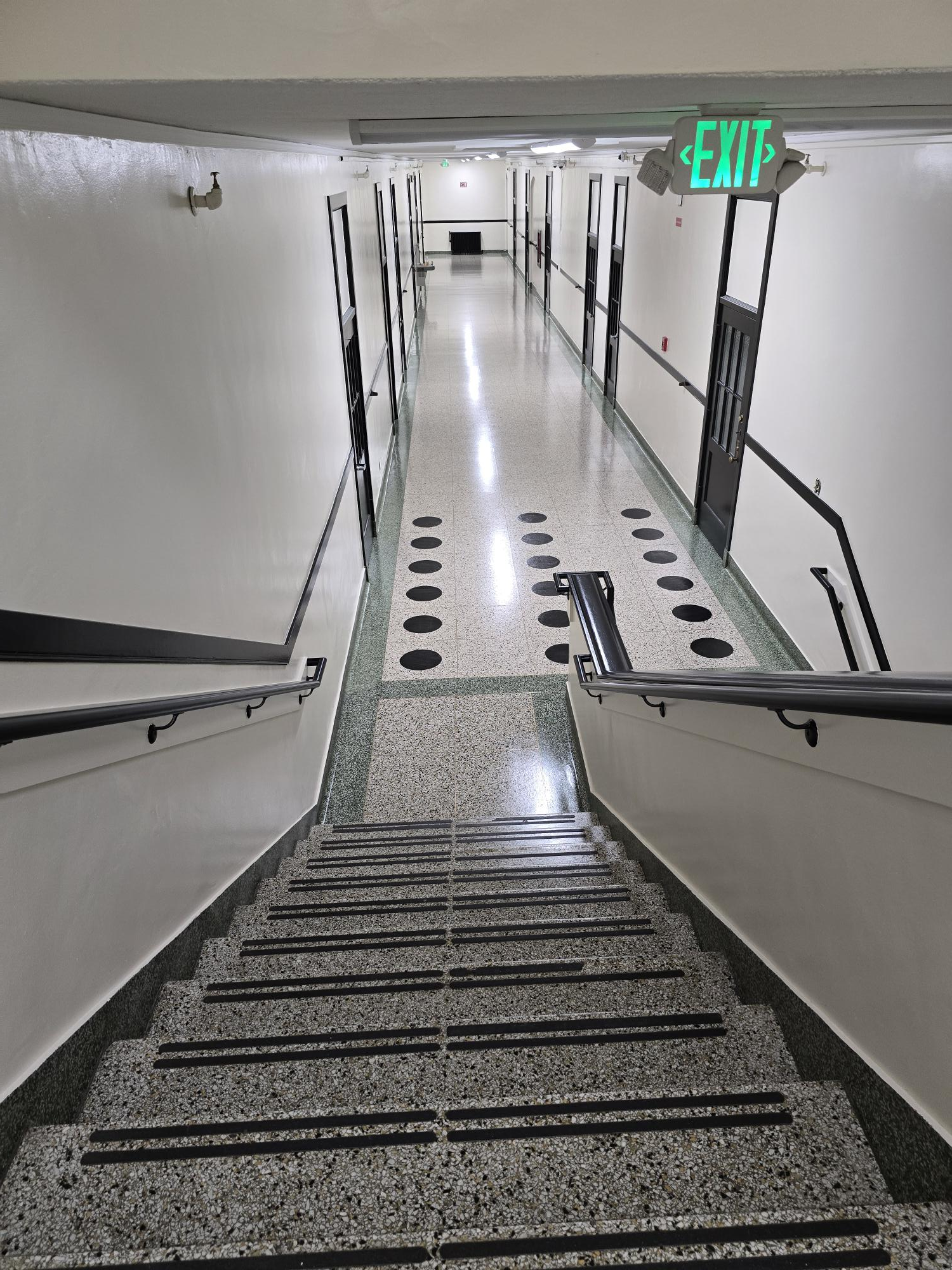 A top-down view of stone stairs leading to a long, sterile white hallway with a glowing green exit sign above.