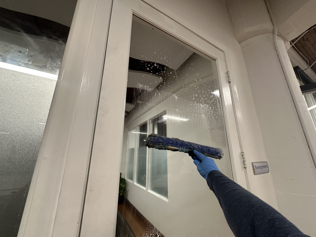 A first-person view of a gloved hand cleaning a glass pane with a blue squeegee in an office hallway.
