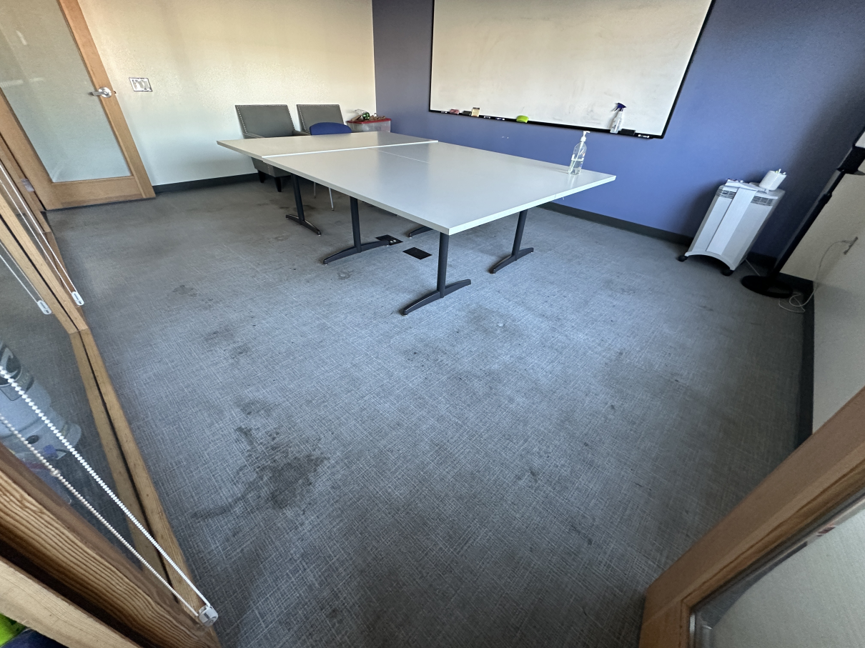 A meeting room featuring a large light-gray table with two chairs, a whiteboard on a blue wall, and a stained gray carpet.