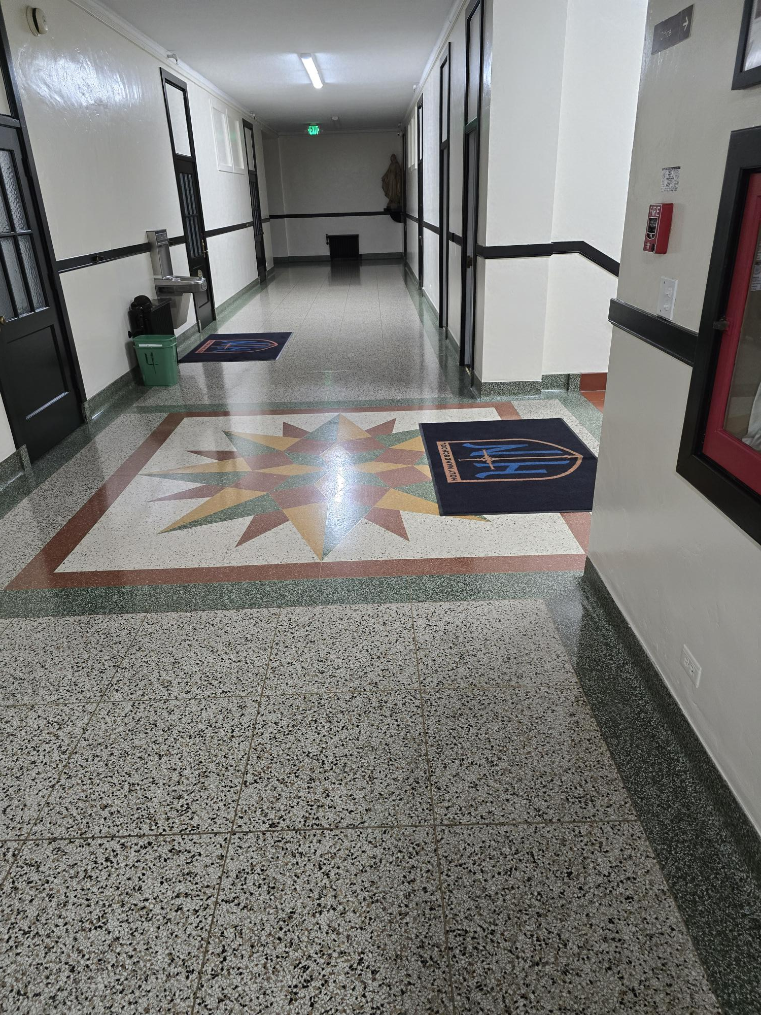 A school hallway with a starburst-patterned floor, dark-framed doors, and two floor mats leading to a distant exit.
