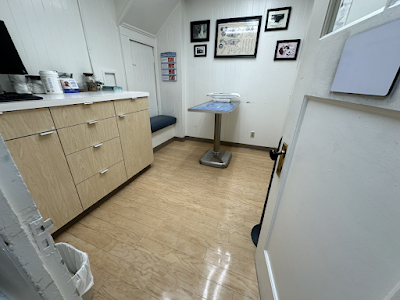 A small medical exam room with light wood cabinets, a stainless steel examination table, and framed wall art.