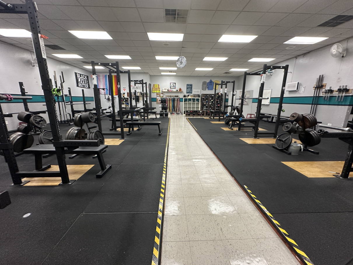 A weightlifting gym with multiple squat racks, benches, and black rubber flooring divided by a central tile walkway.
