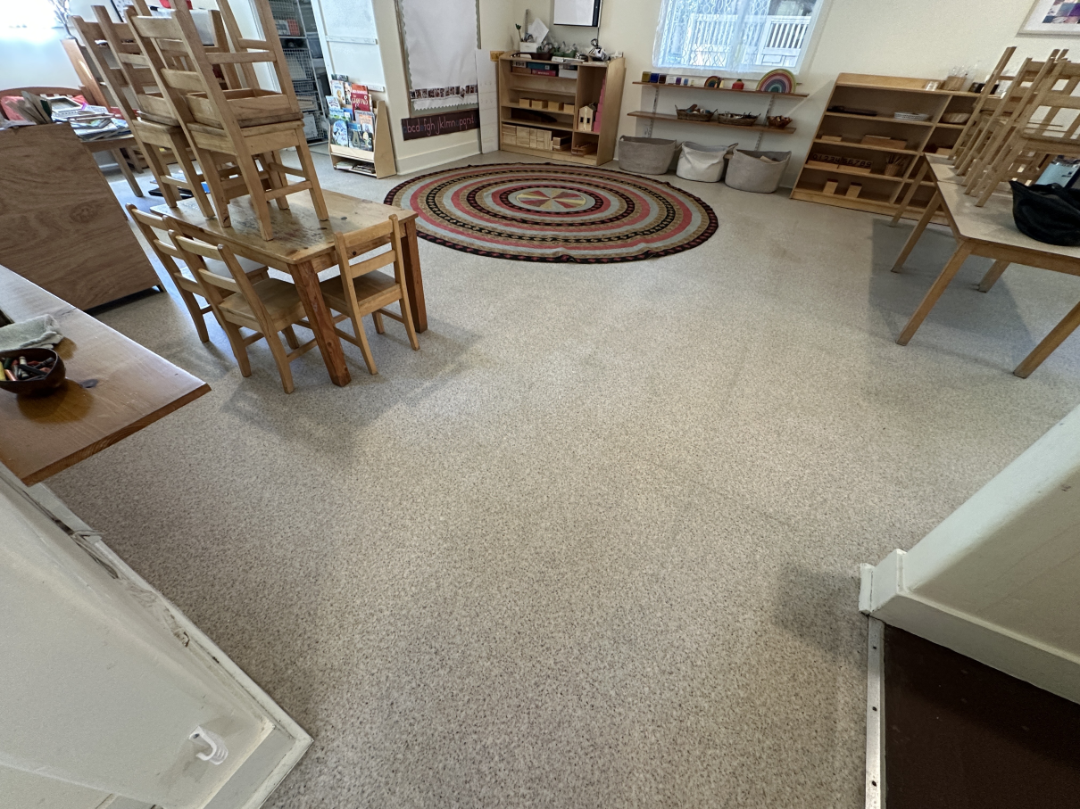 A classroom with wooden tables, small chairs, a round rug, and shelving units filled with learning materials.