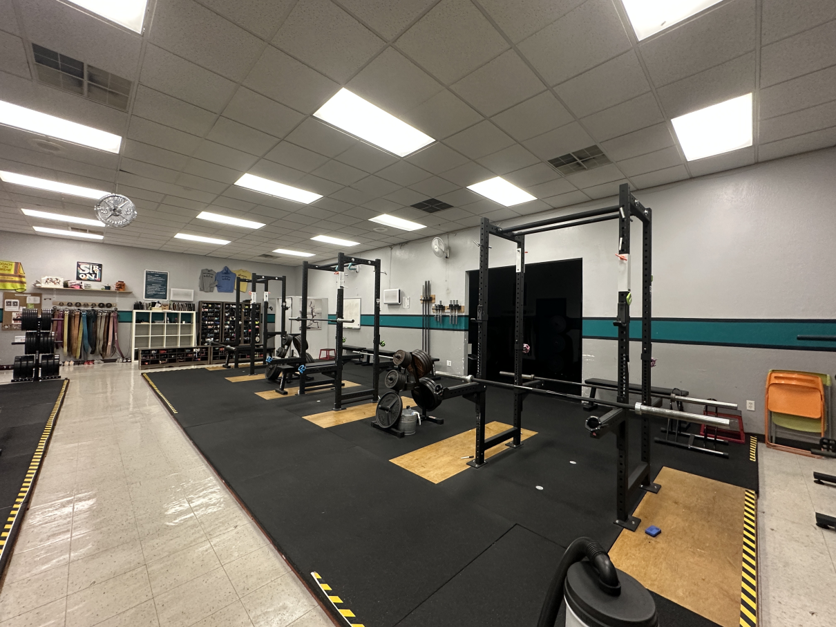 A weight room with several squat racks set up on a large black mat, with a tiled walkway along the left side.