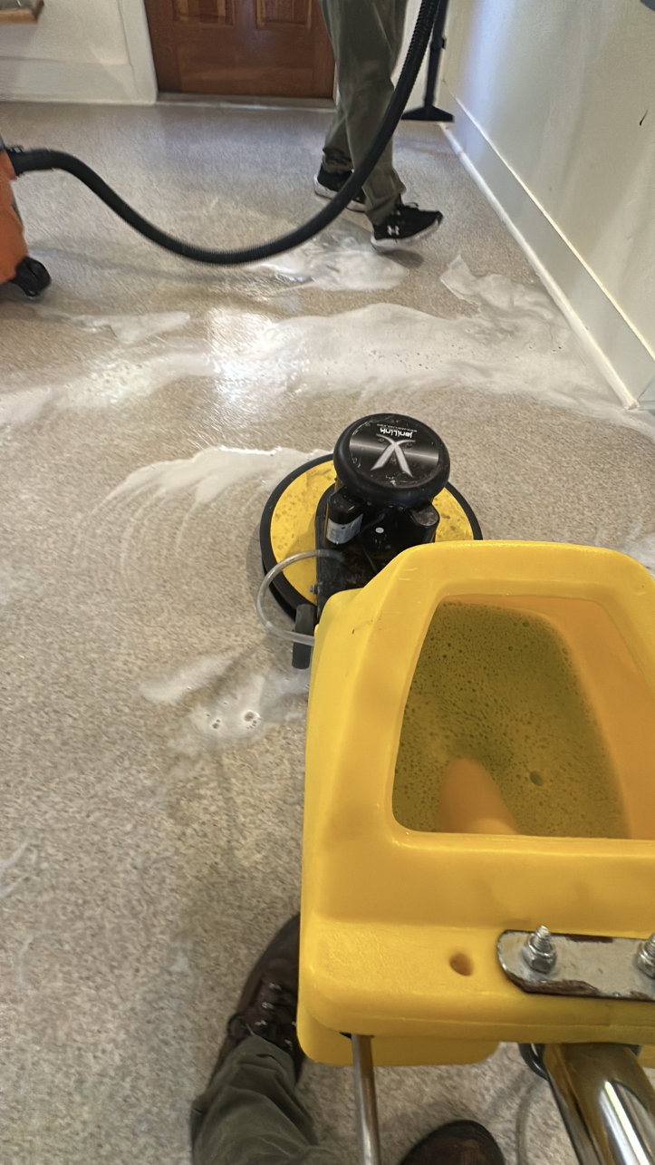 A person operates a floor scrubber on a speckled surface, with another person using a vacuum nearby.