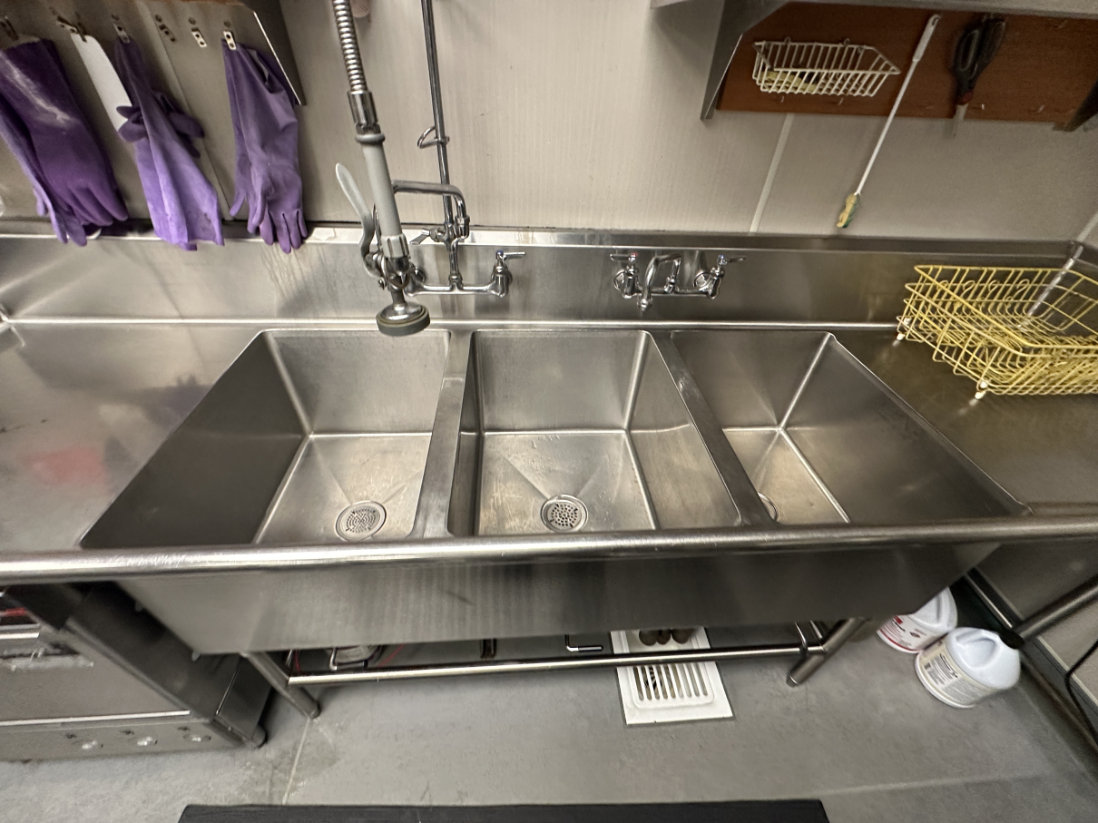A stainless steel, three-compartment commercial sink in a kitchen with purple gloves hanging on the wall.