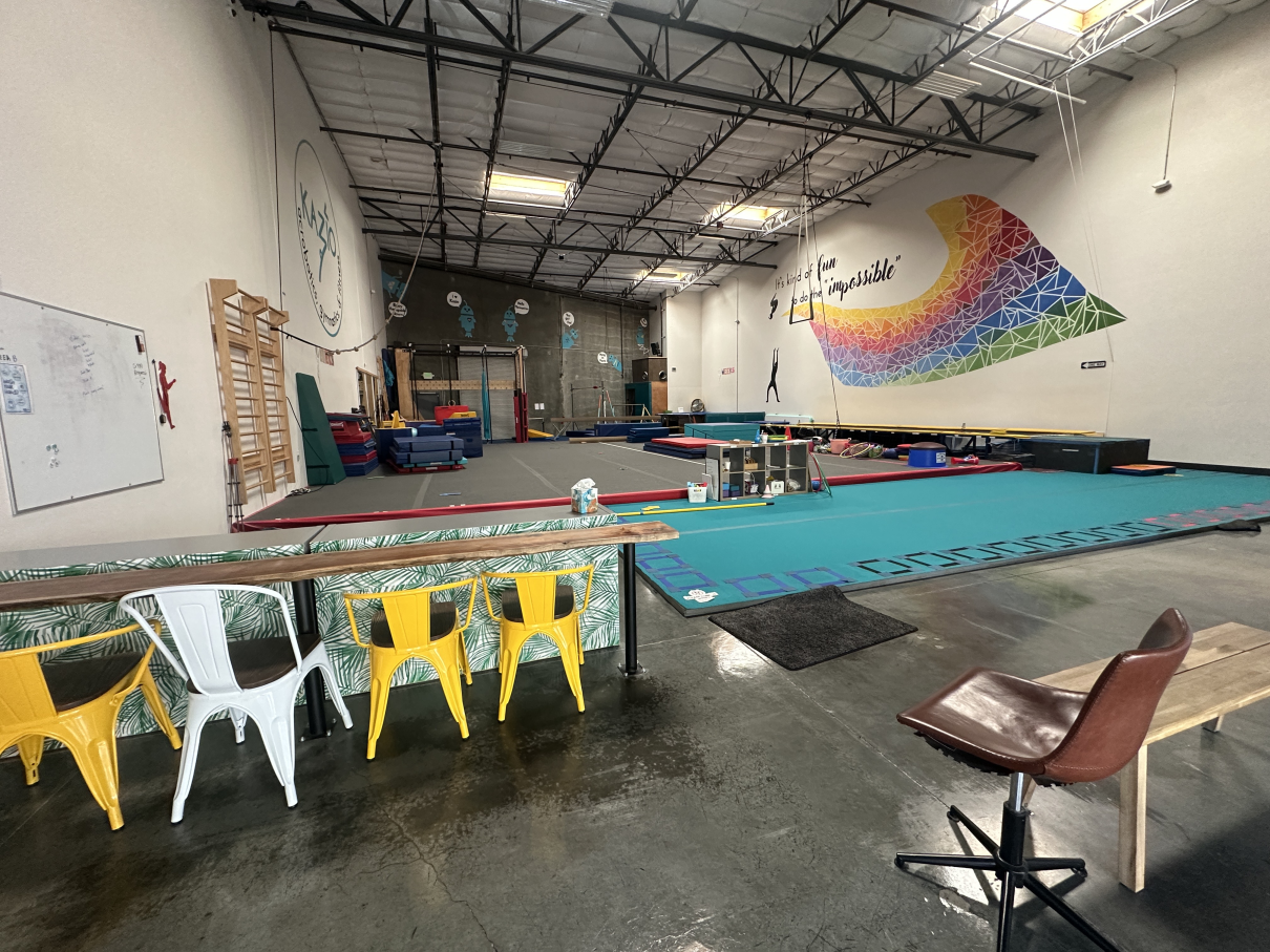 A brightly lit gymnastics studio with a colorful rainbow mural, floor mats, and chairs facing the training area.