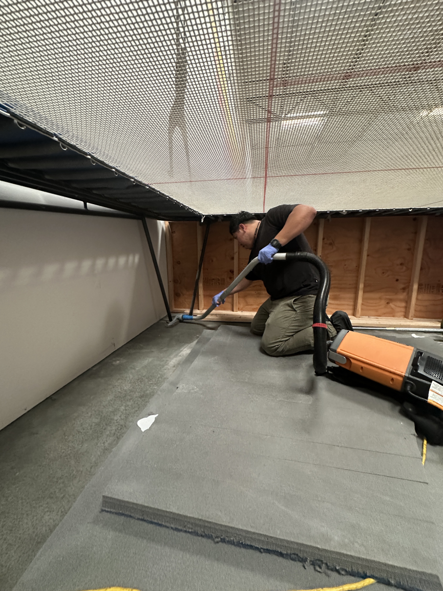 A person wearing gloves and a black shirt kneels on gray carpet, using a vacuum to clean against a wall under a structure.