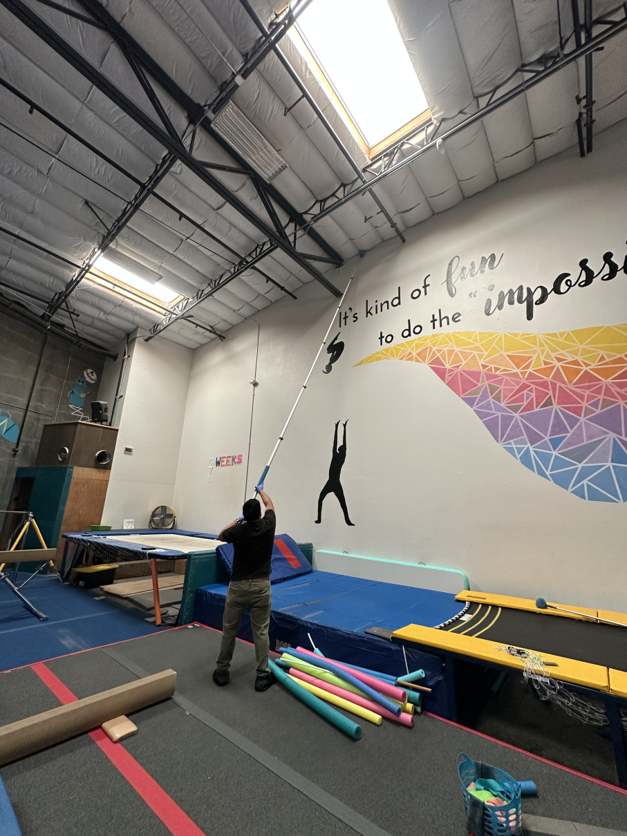 A person uses an extension pole to reach a wall-mounted sensor inside a gym with trampolines and colorful foam mats.