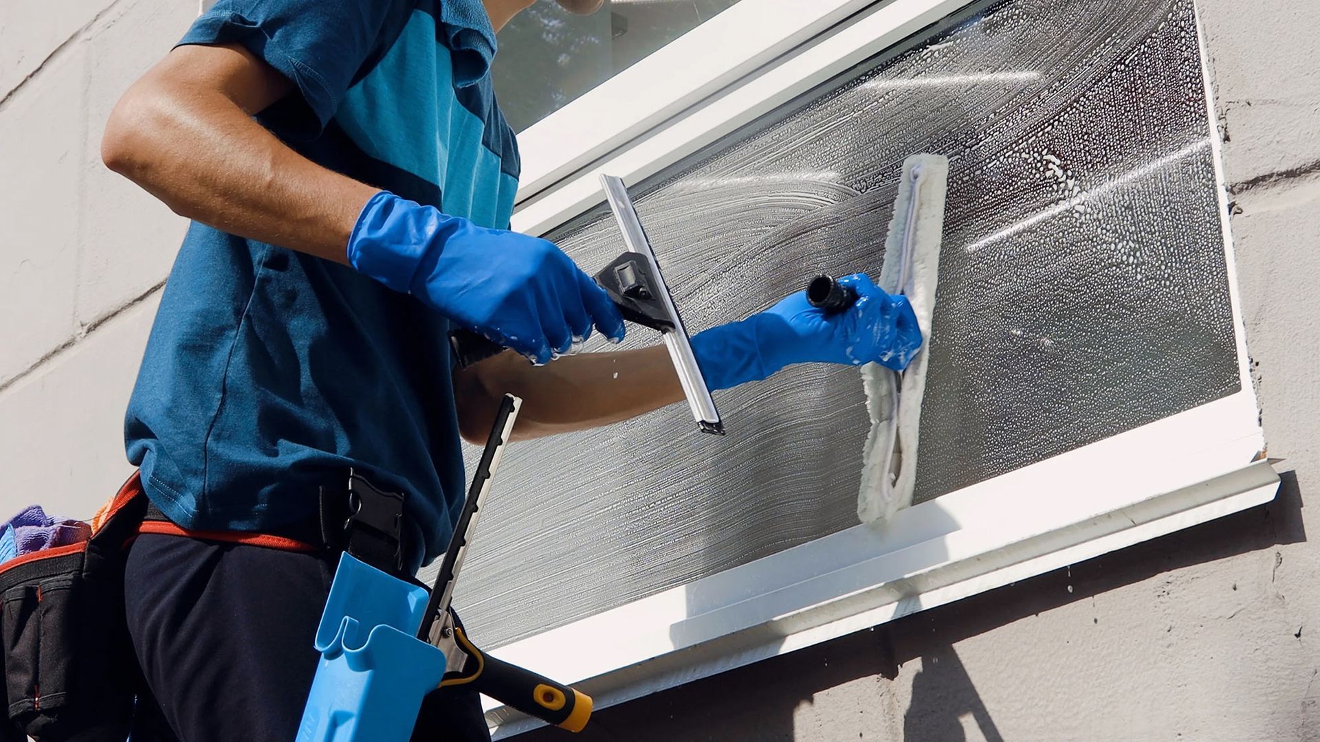 A person wearing a blue glove cleans a glass door with a soapy sponge, leaving water streaks on the surface.