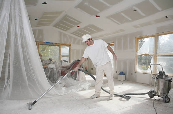 A person uses a wooden-handled bristle brush to sweep construction debris onto a metal dustpan on a white floor.