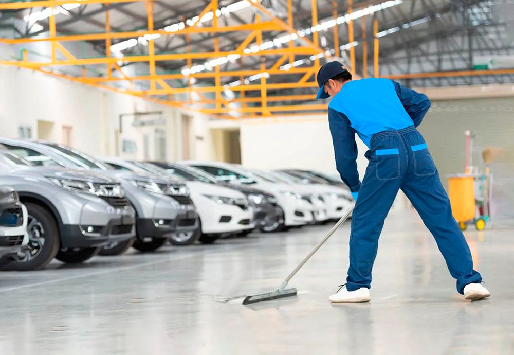 A worker in a blue uniform uses a squeegee to clean the concrete floor of a large facility filled with parked cars.