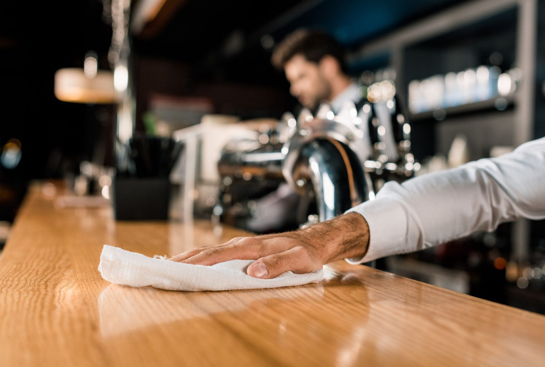 A person in a white shirt wipes down a polished wooden bar top in a dimly lit establishment.