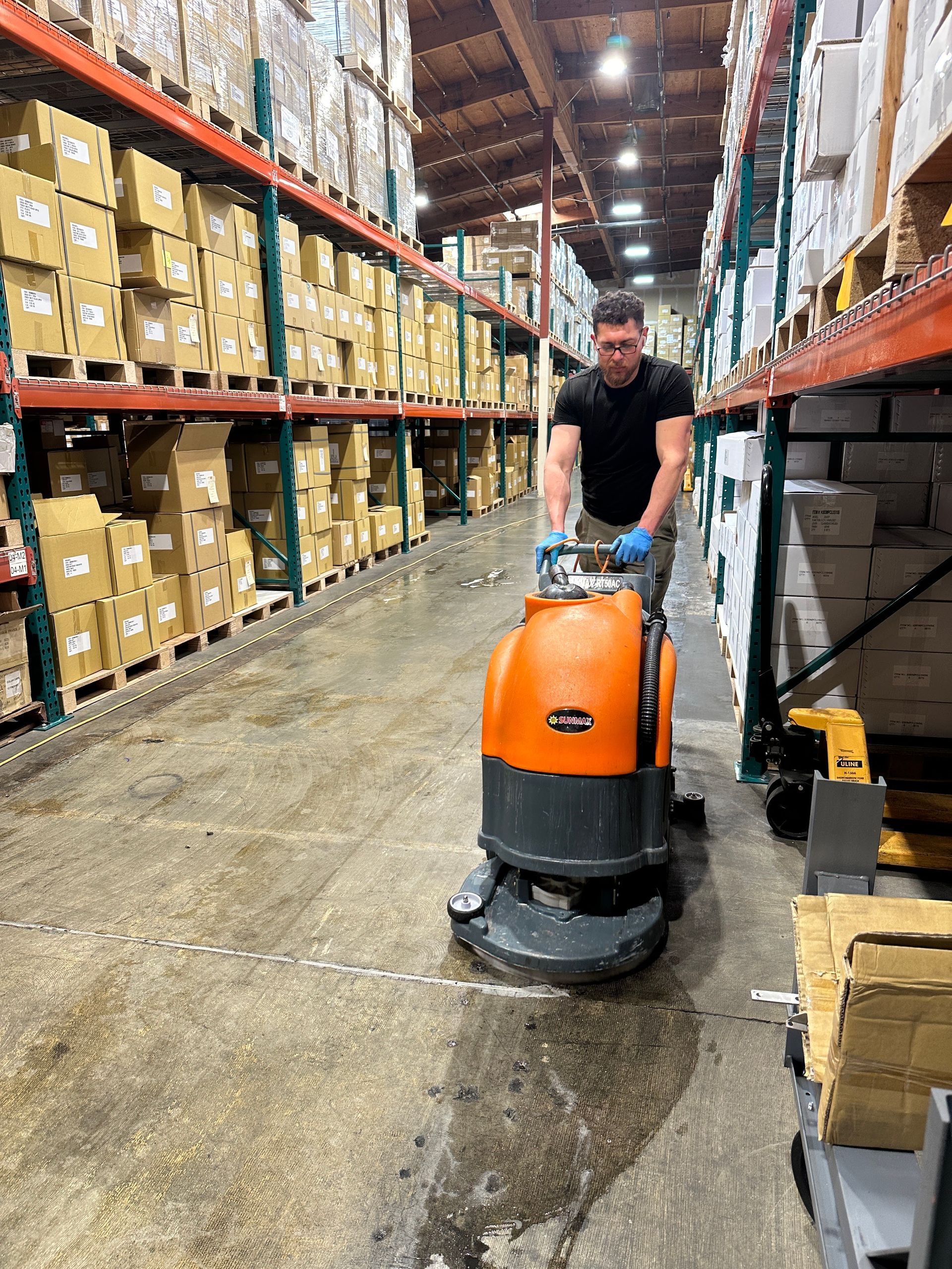 A worker in a black shirt uses an orange industrial floor scrubber in a warehouse with tall aisles of stacked boxes.