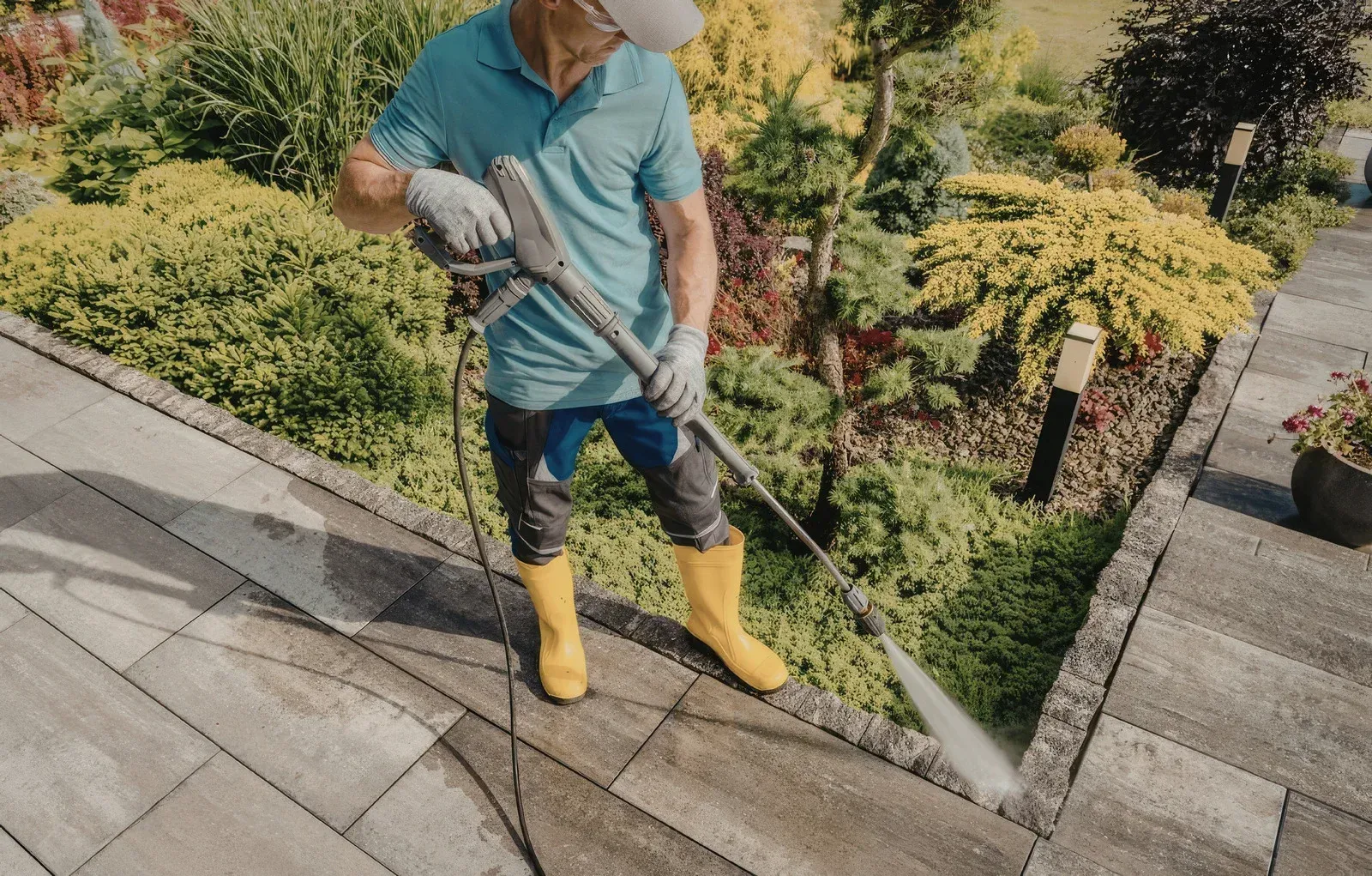 A person in a blue polo shirt and yellow boots uses a pressure washer to clean stone pavers next to a garden.