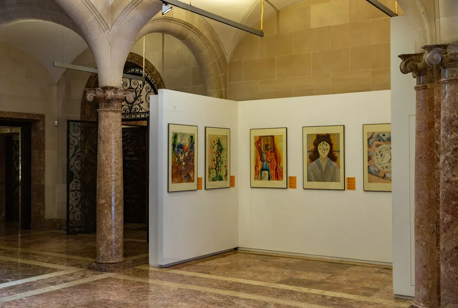 Five framed artworks displayed on a white partition wall inside an ornate hall with stone columns and marble floors.