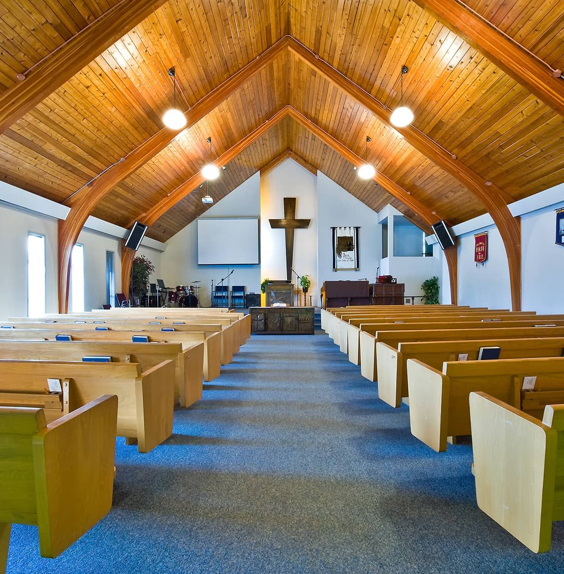 An interior view of a church with wooden vaulted ceilings, rows of wooden pews, and a cross centered at the front.