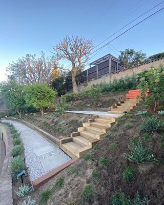 Wooden stairs and paved path ascend a hillside garden, leading to a structure and trees.