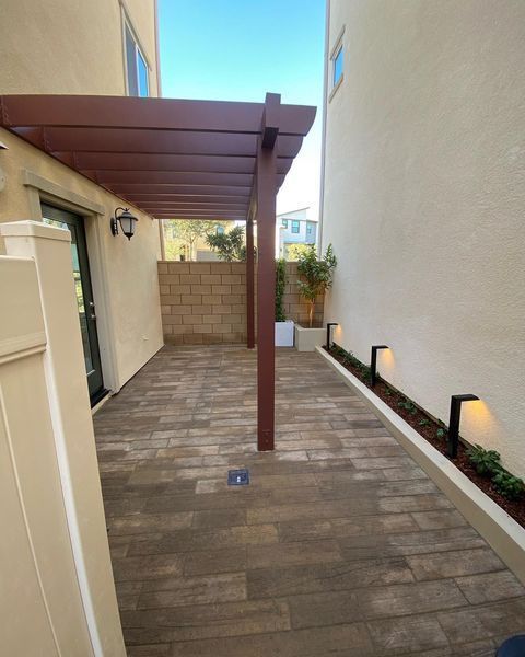 Outdoor patio with pergola, brick pavers, brown pillars, and built-in lighting between beige buildings.