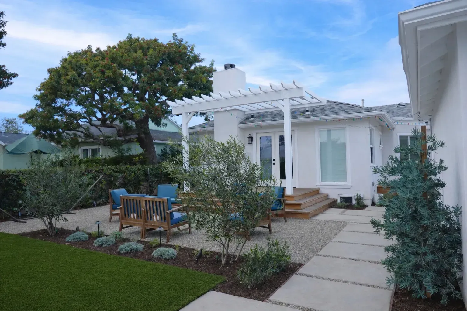 White house patio with pergola, blue chairs, stone path, and landscaped garden under a cloudy sky