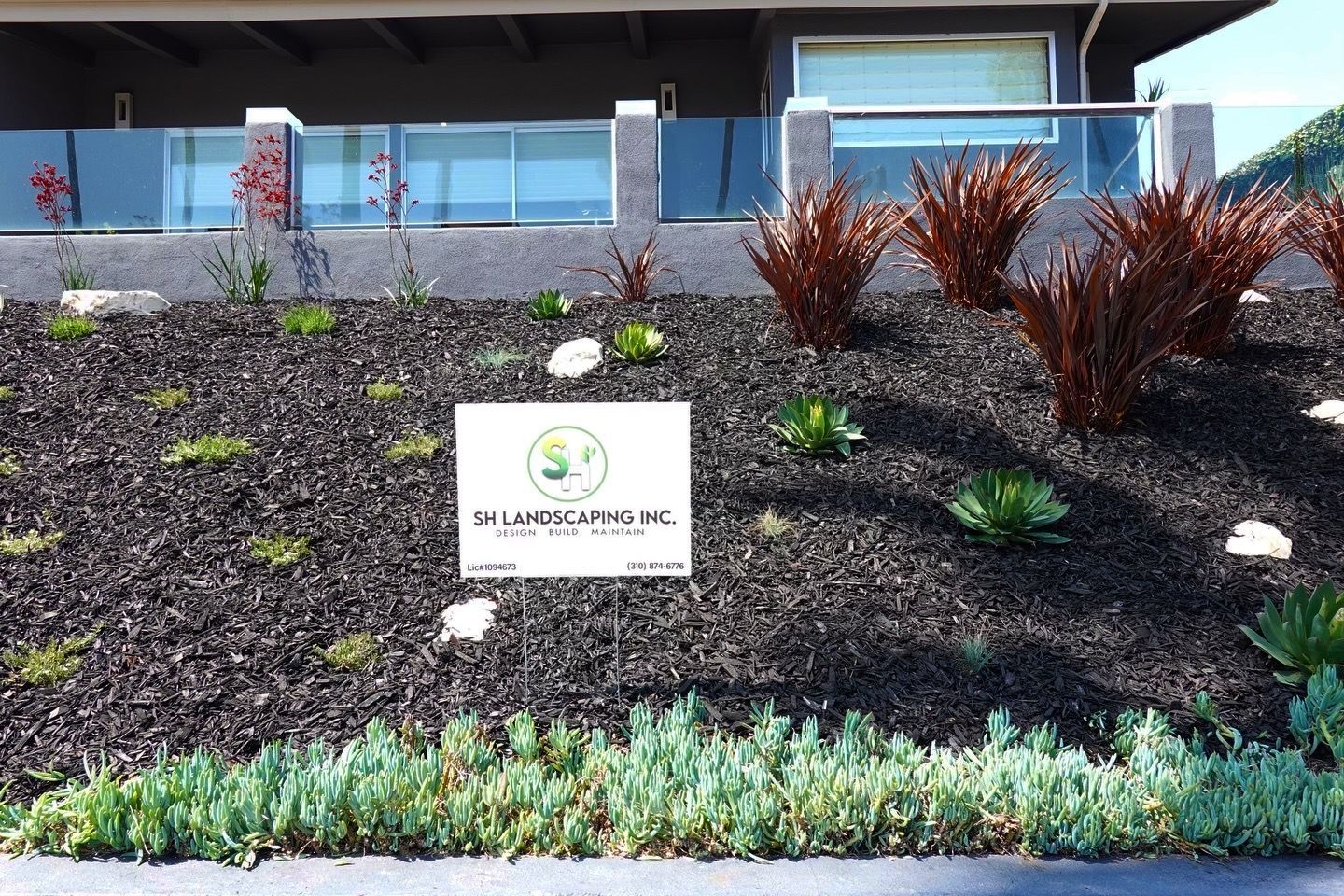 Landscaped yard with dark mulch, red and green plants, and a business sign.