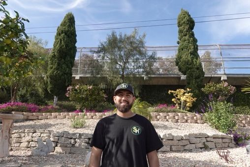 Man in black shirt and hat smiles in front of a garden with stacked stone walls and tall trees.
