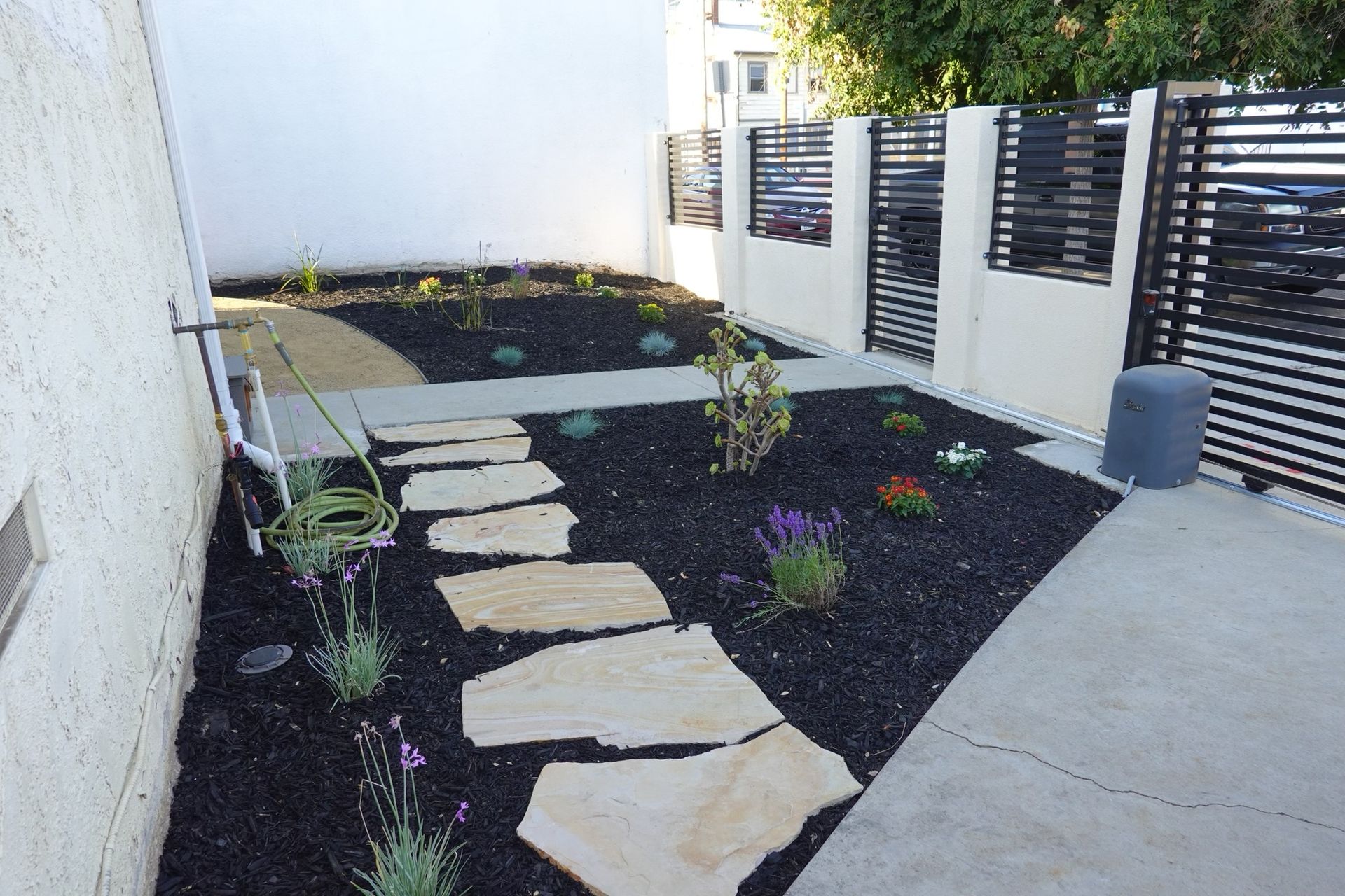 A garden with stone path, black mulch, flowers, and a modern black and beige fence.
