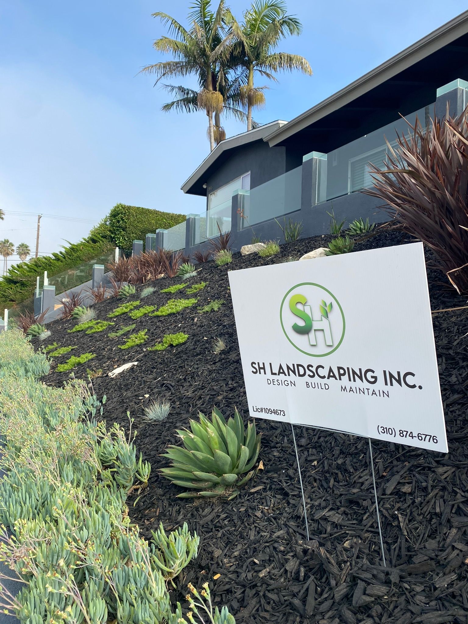 A sign for SH Landscaping Inc. on a landscaped hillside with a house in the background.