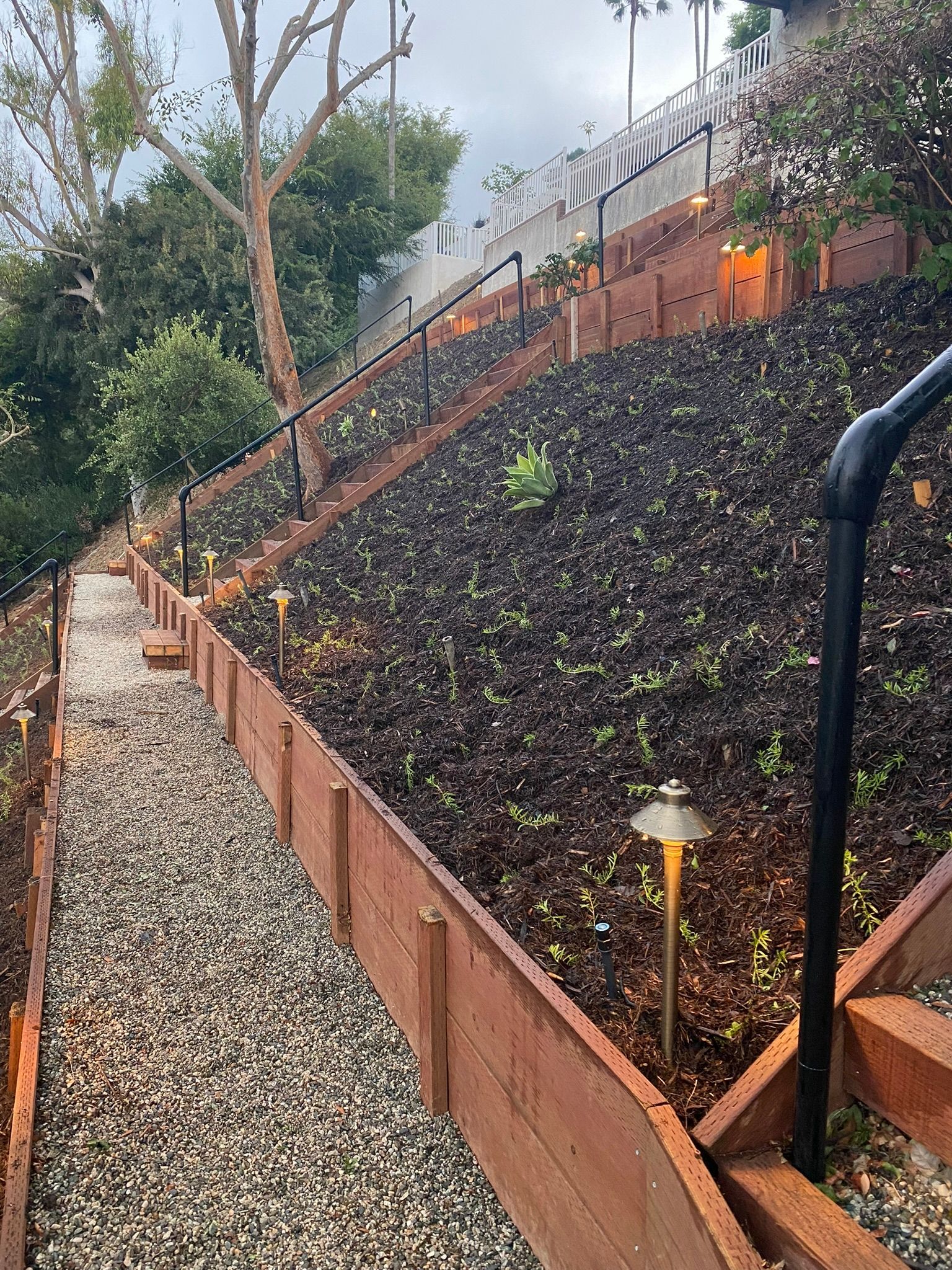 A gravel pathway and mulched slope, with retaining walls, railing, and landscape lighting, ascending a hillside.