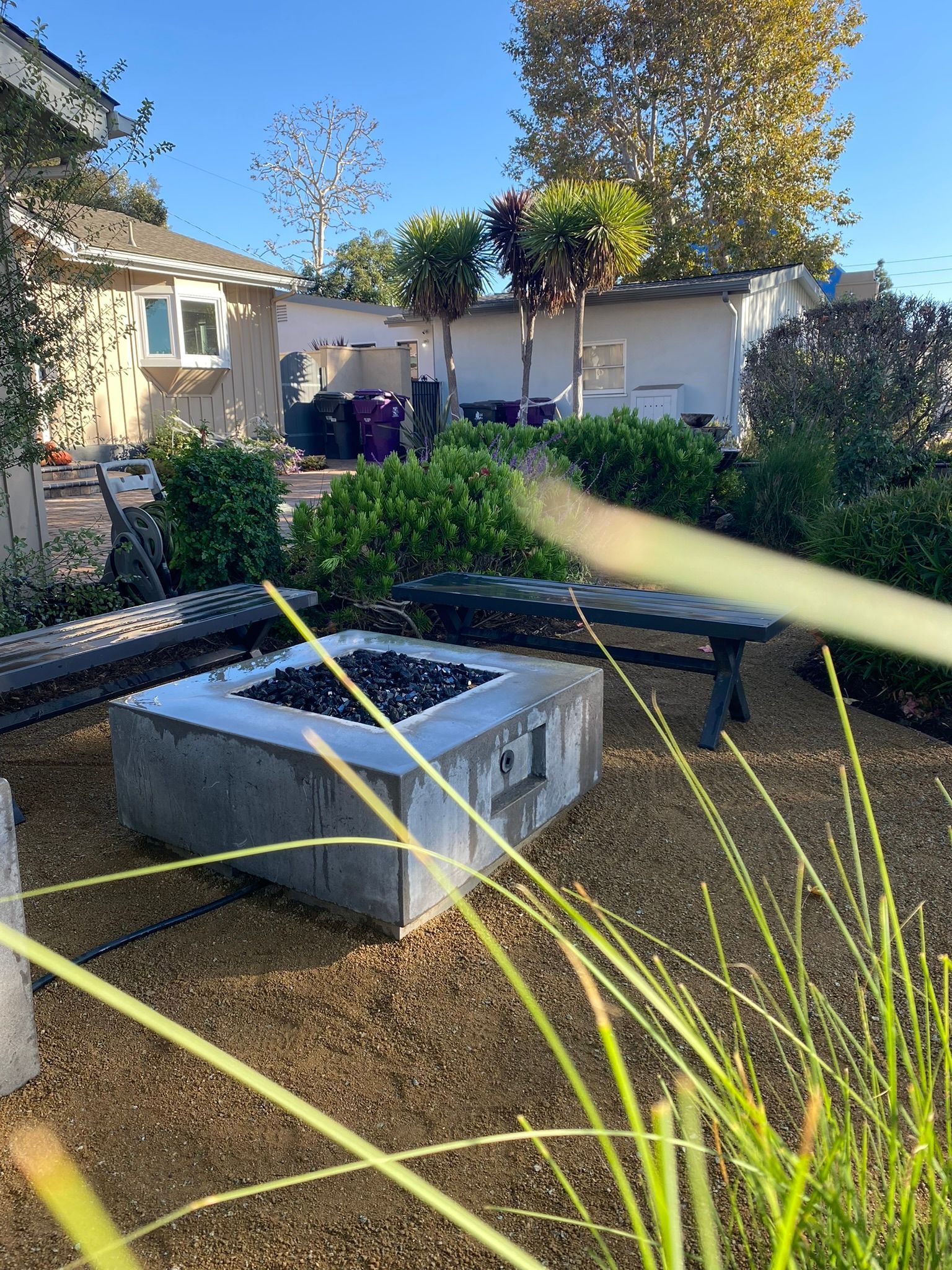 Concrete fire pit in a gravel yard, benches, and trimmed trees.