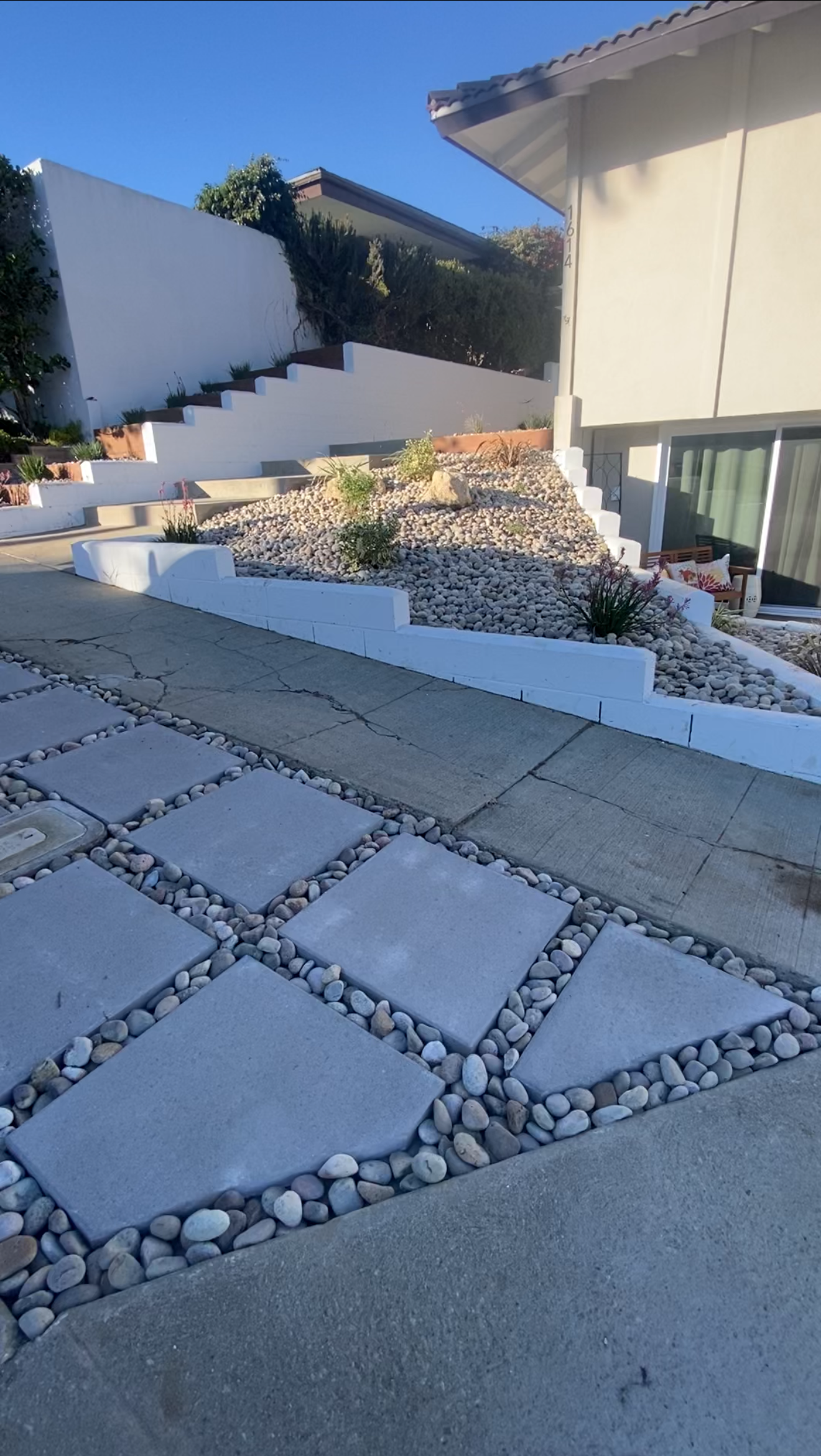 A driveway with geometric pavers bordered by small rocks, leading to a tiered, rock-filled landscape.