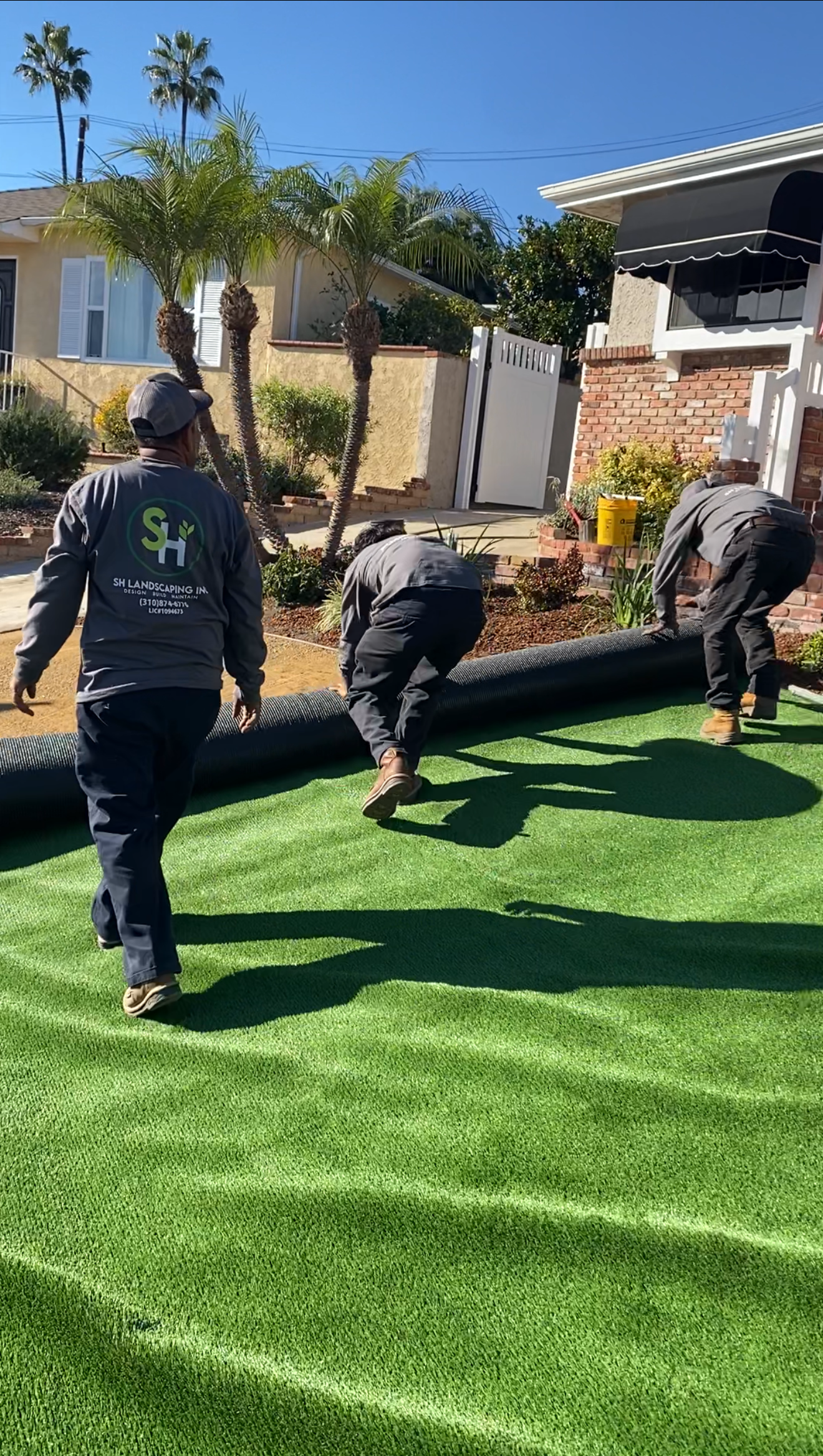 Three workers unrolling artificial turf in a sunny yard; a house and palm trees are visible in the background.