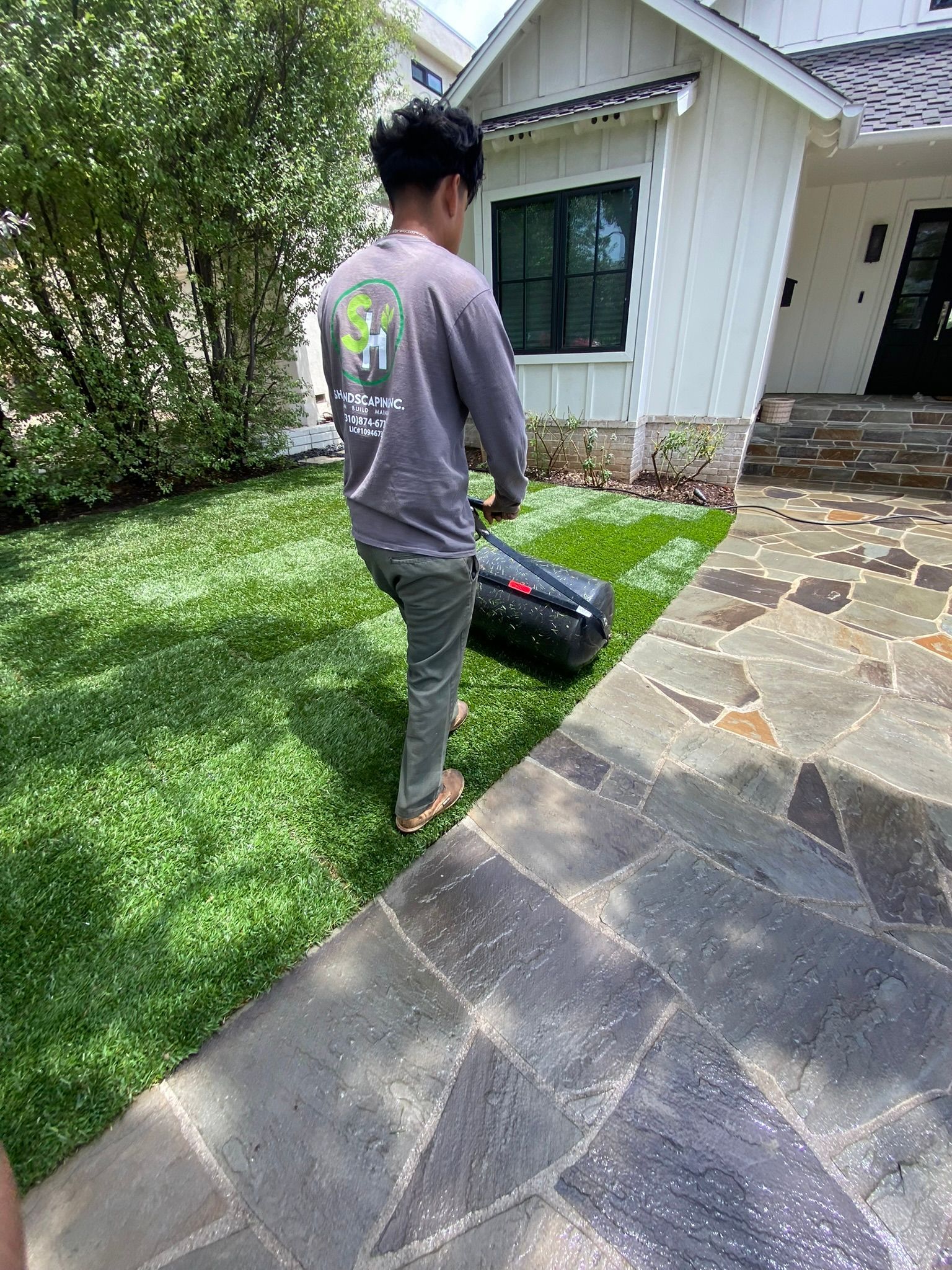 Person in grey shirt and pants pulling a black bag on green grass beside a stone walkway.