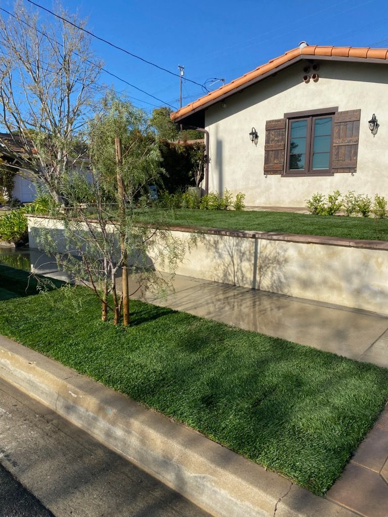 Green lawn and tree in front of a stucco house with a red tile roof and brown shutters on a sunny day.