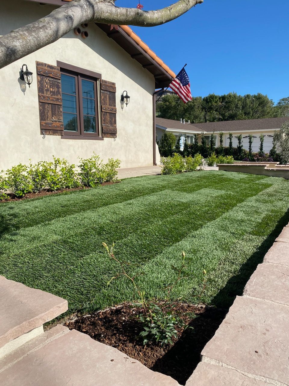 Lawn with alternating dark and light stripes in front of a tan house with a US flag.