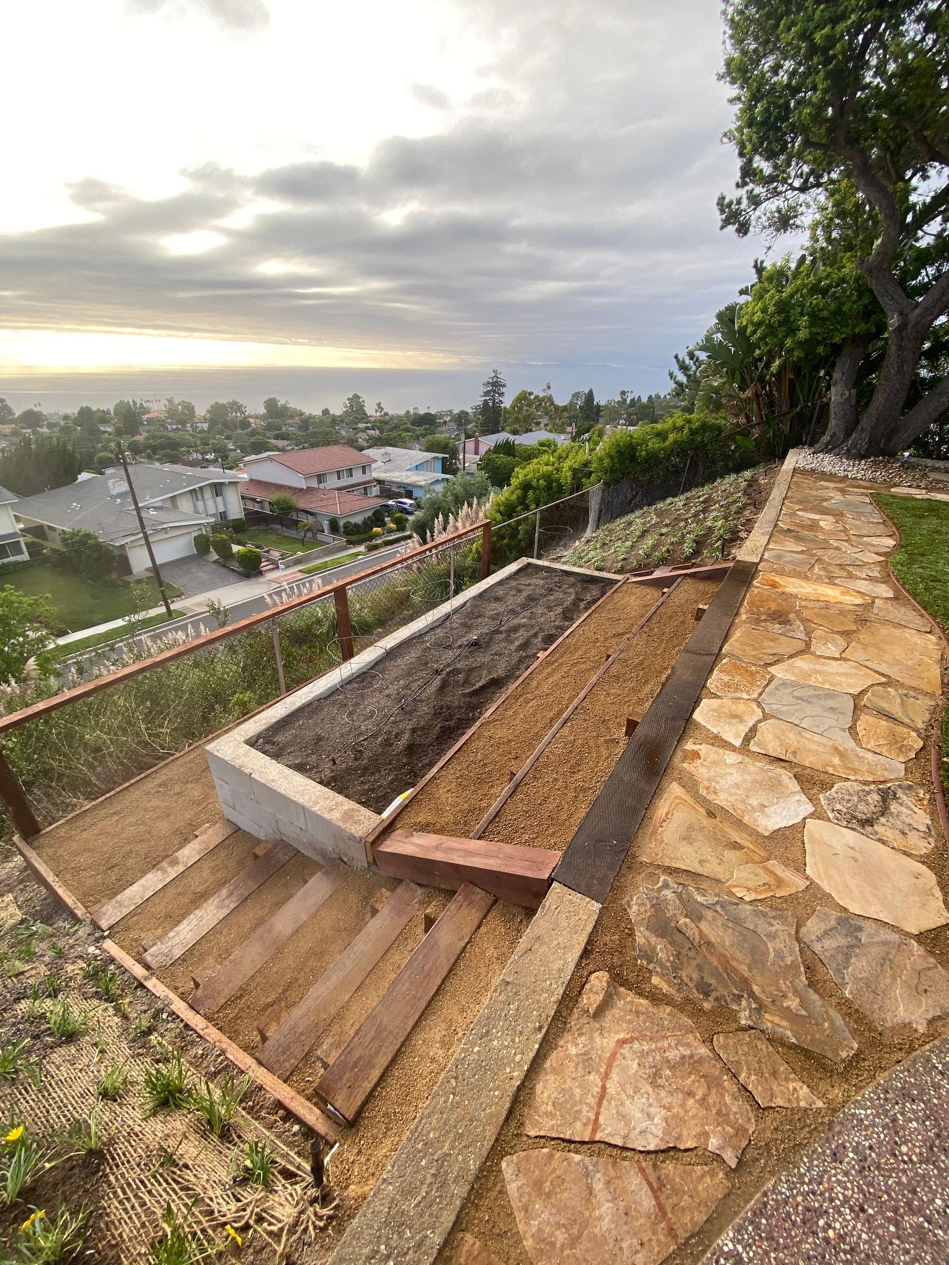 Raised garden beds with view of ocean, city. One empty, one filled with dirt. Stone pathway. Cloudy sky.