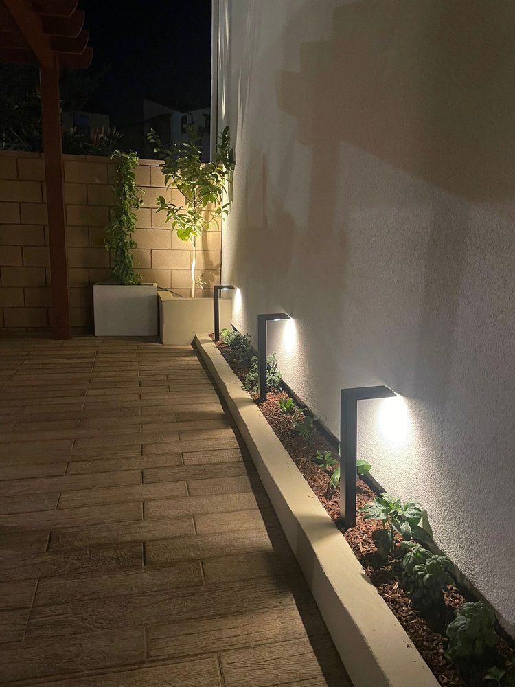 Pathway lit by modern lights along a wall, with a stone path and small plants in a garden bed.
