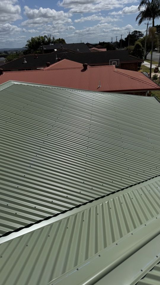 A high-angle view of a sage green corrugated metal roof with a neighboring red metal roof under a partly cloudy sky.