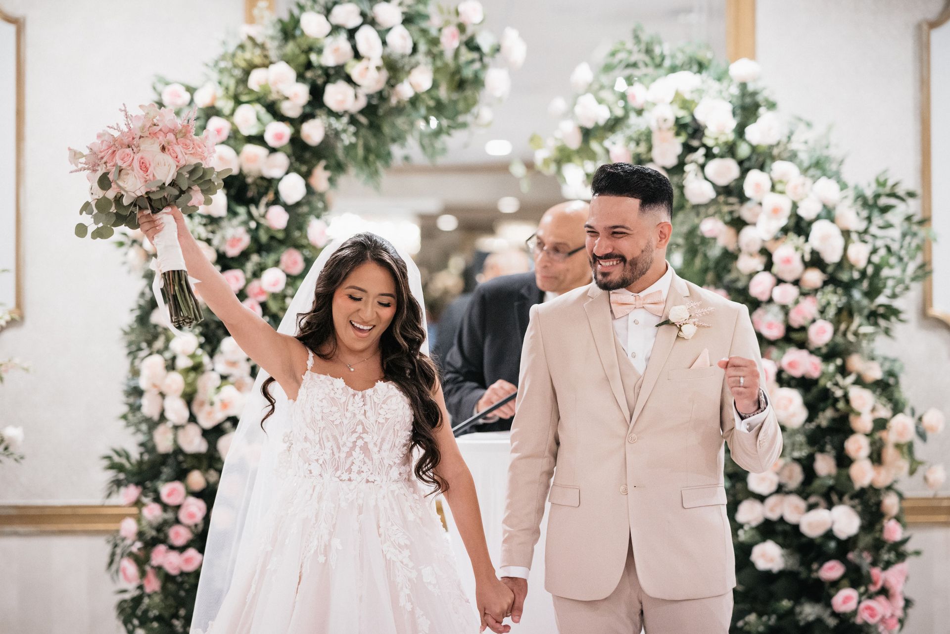 A bride and groom are walking down the aisle holding hands. floral arch by weddings by maritzas 