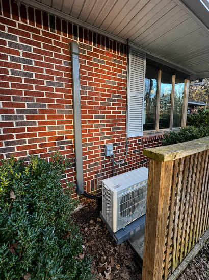 Brick exterior wall with utility pipes, an air conditioner unit, and bushes beside a window and porch.