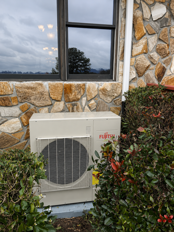 Outdoor air conditioning unit beside a stone wall and window, partly hidden by shrubs